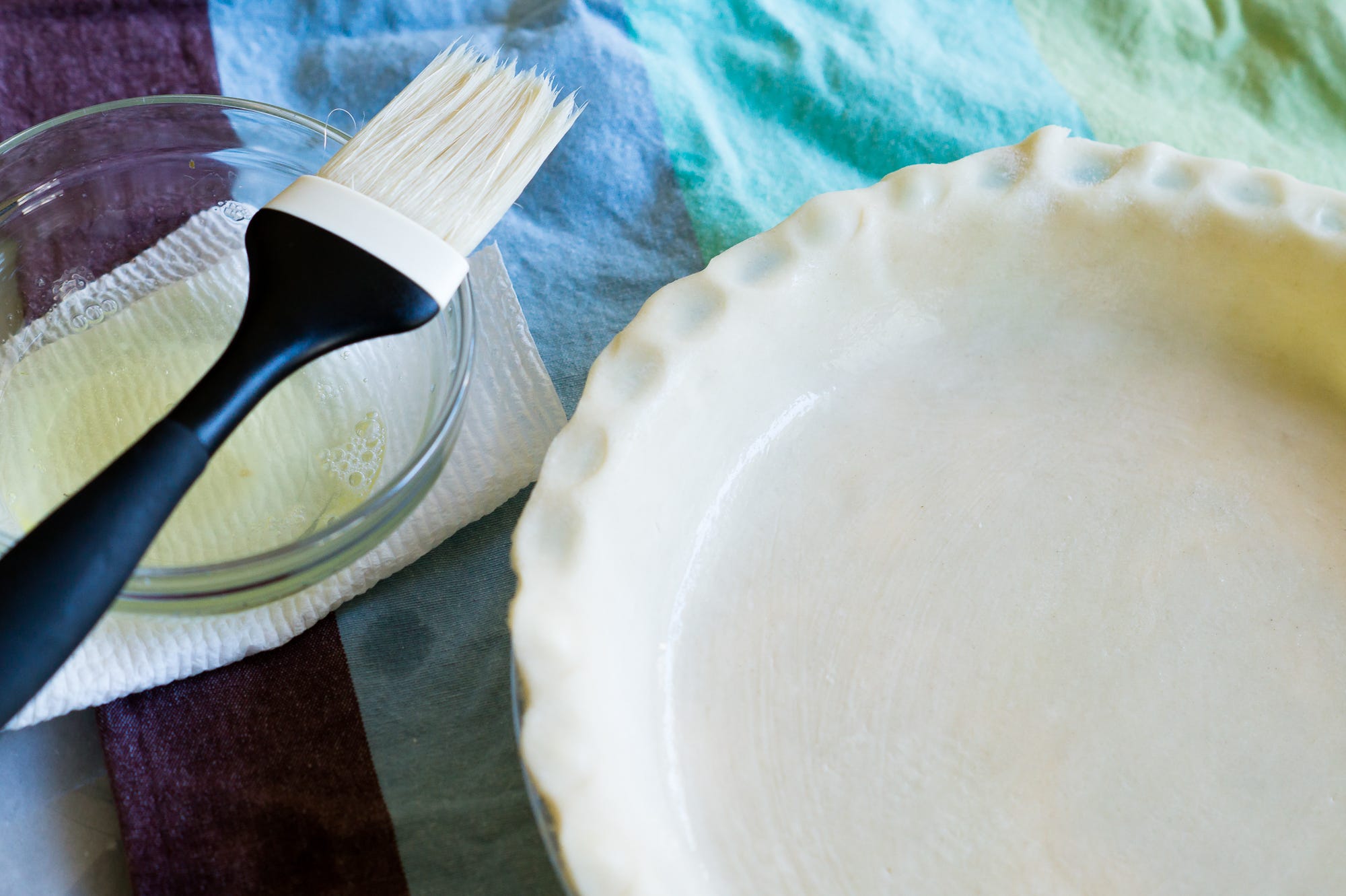 A pastry brush resting on a bowl of egg wash beside an unbaked pie crust, warm kitchen tones